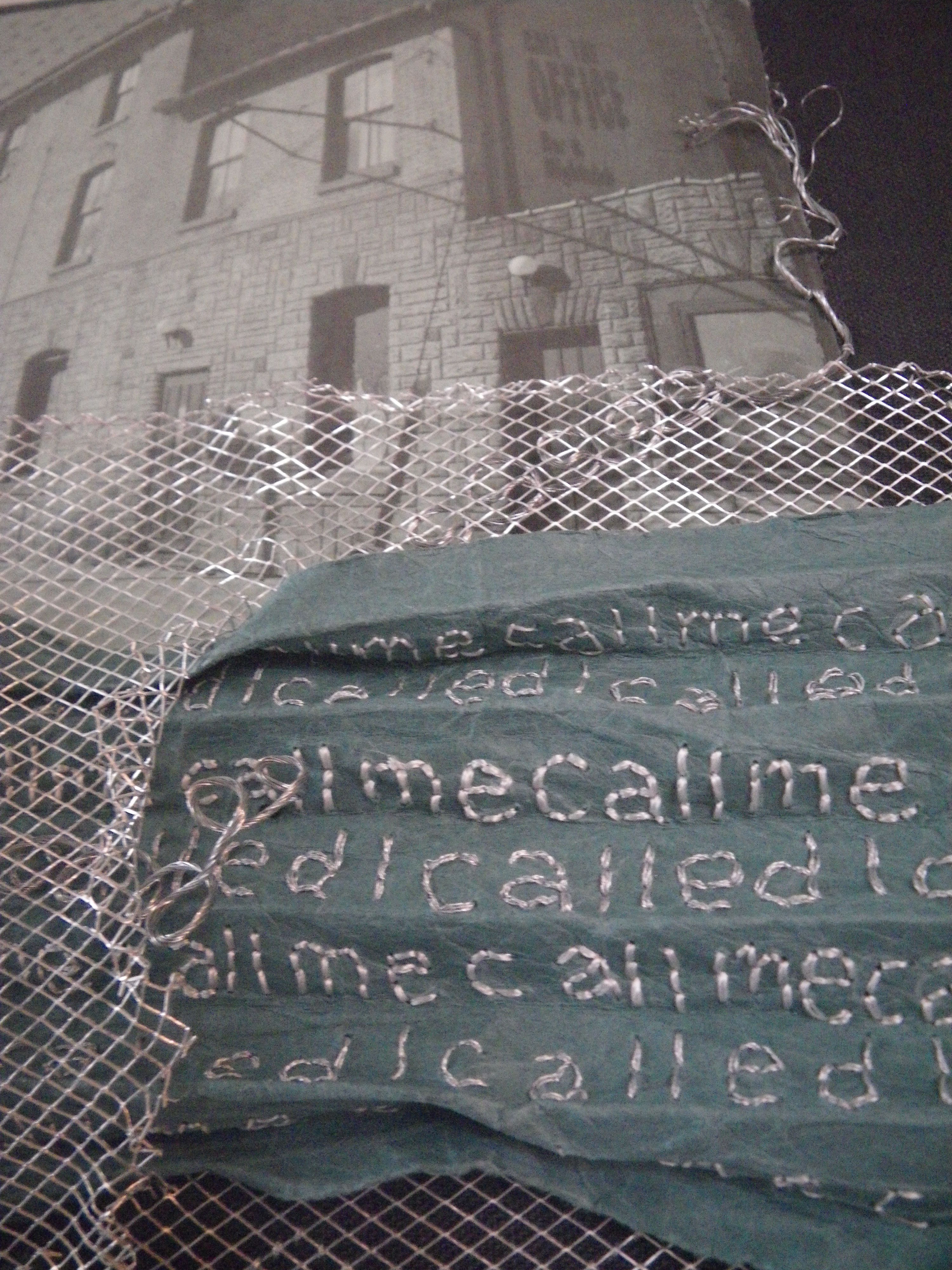 detail of Missed Call, paper coming though silver mesh fencing; a bar "Call the Office" in the background